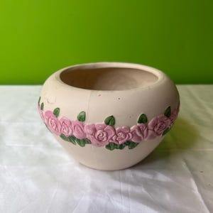 May include: A white ceramic planter with a pink rose design. The planter has a rounded shape and a wide opening.