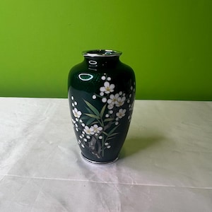 Puede incluir: Un jarrón verde oscuro con un borde plateado y un diseño floral de flores blancas y hojas verdes.