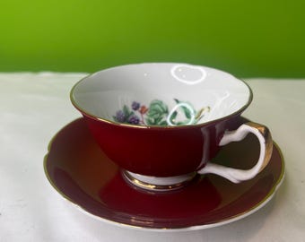 May include: A red teacup and saucer set with a floral design. The cup and saucer have a gold trim.