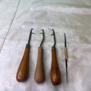 May include: Three crochet hooks with wooden handles and a single needle tool. The hooks have metal tips and are labeled "MADE IN KOREA" and "MADE IN USA". The tools are arranged on a white surface.