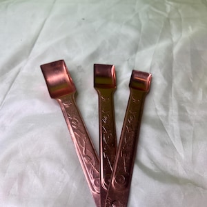May include: Three copper-colored ice tongs with a rectangular scoop end and a decorative etched design on the handle. The tongs are arranged in a fan shape, showcasing their intricate details and warm metallic hue.