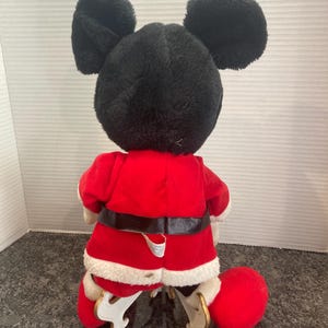 May include: A plush Mickey Mouse doll wearing a red Santa Claus jacket with white faux fur trim and a black belt. The doll has large black ears and is positioned on white plastic skates. The back of the doll is visible, showing the red fabric.