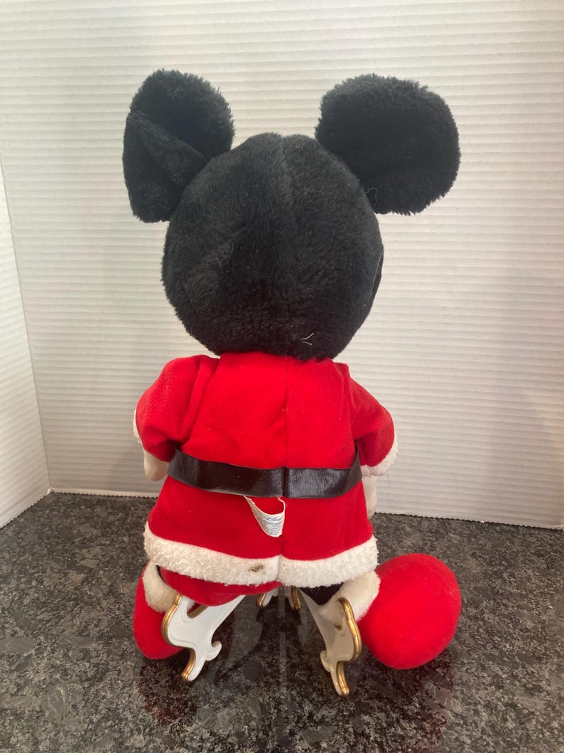 May include: A plush Mickey Mouse doll wearing a red Santa Claus jacket with white faux fur trim and a black belt. The doll has large black ears and is positioned on white plastic skates. The back of the doll is visible, showing the red fabric.