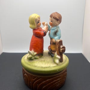 May include: A vintage porcelain music box featuring a boy and girl figurine holding a white dove. The music box is decorated with green grass and flowers and has a brown wooden base.