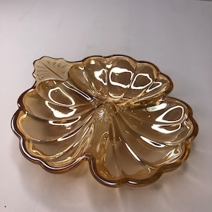 Puede incluir: Un plato de vidrio ámbar claro con forma de flor y un detalle de hoja. El plato tiene un borde festoneado y es perfecto para servir aperitivos o pequeñas golosinas.