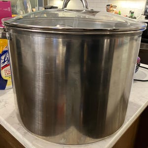Puede incluir: Una olla grande de acero inoxidable con tapa de cristal transparente y asa plateada. La olla es cilíndrica y está sobre una encimera blanca. La olla está diseñada para cocinar grandes cantidades de alimentos.