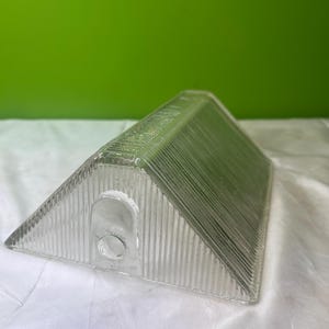 Puede incluir: Una pantalla de lámpara triangular de vidrio transparente con detalles acanalados verticales. La pantalla tiene una pequeña abertura circular en un lado. El fondo es verde.