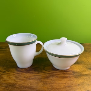 Puede incluir: Un juego de crema y azucarero blanco. Ambas piezas tienen una banda verde con un borde dorado. La crema tiene un asa y un pico. El azucarero tiene una tapa con un pequeño pomo.