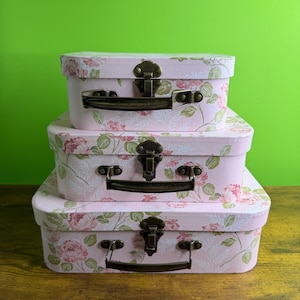 May include: Three stacked, oval-shaped decorative boxes. The boxes are light pink with a floral pattern of pink roses and green leaves. Each box has a dark bronze-colored handle and clasp. The boxes are on a wooden surface with a green background.