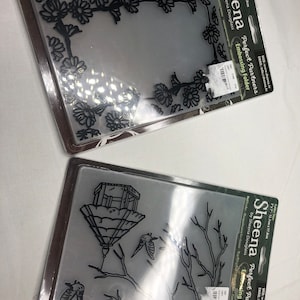 Peut inclure: Deux pochettes à embosser en plastique transparent avec des motifs noirs. La première pochette présente une bordure florale avec un cadre rectangulaire au centre. La deuxième pochette présente un nichoir à oiseaux avec des oiseaux volant autour d'une branche d'arbre. Les deux pochettes sont étiquetées "Sheena Perfect Partners Embossing Folder" et incluent le site Web et l'adresse e-mail de Sheena Douglass.
