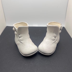 Könnte beinhalten: Ein Paar weiße Keramik-Stiefel mit Knopfdetails. Die Stiefel sind klein und haben eine abgerundete Spitze.