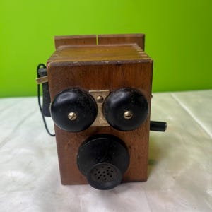 Puede incluir: Un teléfono de madera antiguo con campanas circulares negras y una boquilla. El teléfono tiene un cuerpo rectangular de madera con un acabado oscuro y detalles en latón. Se ven un cable negro y una pequeña palanca.
