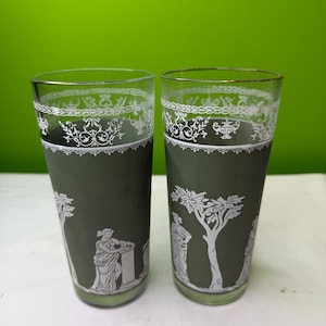 May include: Two vintage green-tinted drinking glasses with white decorative patterns. The glasses feature a white band with ornate designs near the rim and classical figures and trees on the body. The rims have a gold trim.