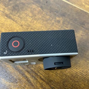 May include: A black action camera with a silver frame. The camera has a black lens and a red circle button. The top of the camera is textured with a diamond pattern.