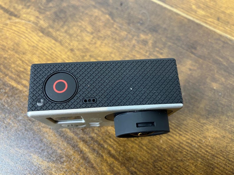 May include: A black action camera with a silver frame. The camera has a black lens and a red circle button. The top of the camera is textured with a diamond pattern.
