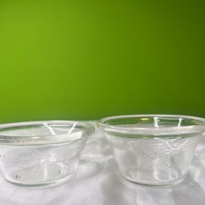 May include: Two clear glass bowls with a decorative floral pattern. The bowls are round with a slightly flared rim. The bowls are on a white surface with a green background.