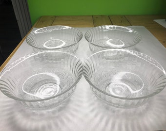 May include: Four clear glass bowls with a decorative pattern. The bowls are arranged in a square shape on a white surface.