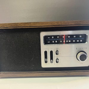 Pode incluir: Um rádio Zenith vintage com uma moldura de madeira escura e um painel de controle prateado. O rádio possui um dial de sintonia, controles de volume e um alto-falante coberto com tecido preto. O logotipo da Zenith é visível.