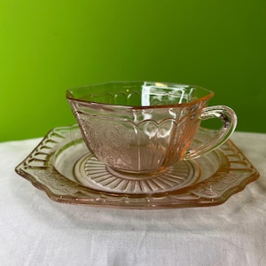 Könnte beinhalten: Ein rosafarbenes Glas-Tee-Set mit einem Blumenmuster. Tasse und Untertasse haben einen gewellten Rand.