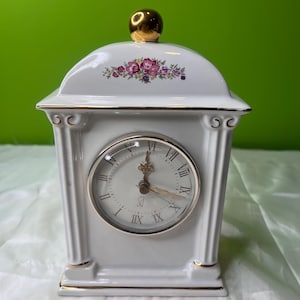 May include: White porcelain mantel clock with gold accents. The clock features a round face with Roman numerals and a decorative floral design on the top. The clock has a gold-colored knob on top.