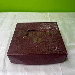 Puede incluir: Una caja color burdeos con una etiqueta dorada que dice "Rechessy" y un logotipo dorado con las letras "SR".