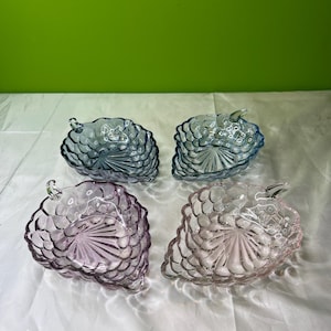 Op de afbeelding: Vier glazen serveerschalen in de vorm van druiventrossen. De schalen zijn in tinten blauw, paars en roze. Elke schaal heeft een textuuroppervlak en een klein handvat. De schalen staan op een wit oppervlak.