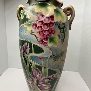 Może przedstawiać: Wysoki, zielony ceramiczny wazon z dwoma uchwytami. Wazon jest ozdobiony kwiatowym wzorem z fioletowych irysów i zielonych liści. Na wazonie namalowano również winogrona i winorośl.