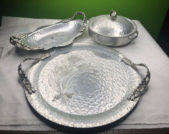 May include: A set of three silver hammered aluminum serving trays with ornate handles. The trays are oval, round, and round with a lid.