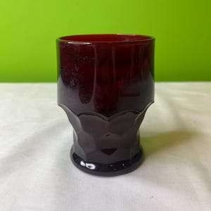 May include: A dark red glass tumbler with a textured, faceted design. The glass is clear and has a slight purple hue.