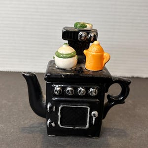 May include: A black ceramic teapot shaped like a stove, with a small white and green lidded pot, and an orange teapot on top. The stove has silver burner details and a white door.