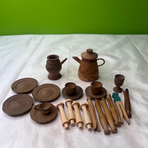 Puede incluir: Una colección de artículos de cocina de madera en miniatura, que incluye platos, tazas, platillos, una tetera, una jarra, rodillos y pinzas para la ropa. Los artículos son de varios tonos de marrón y beige.
