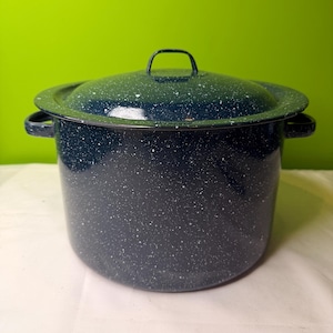May include: A large, dark blue enamel pot with a matching lid. The pot has a speckled pattern of white dots and two side handles. The lid has a small handle on top. The pot is sitting on a white surface with a green background.