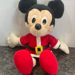 May include: A plush Mickey Mouse toy dressed in a red Santa Claus outfit with white trim, a black belt, and a yellow square buckle. The character has large black ears, a tan face, white gloves, and red shoes, sitting in a forward-facing position.