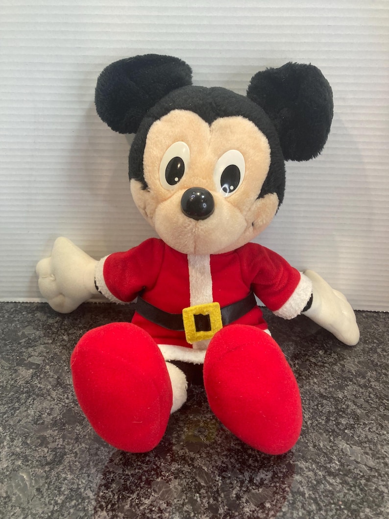 May include: A plush Mickey Mouse toy dressed in a red Santa Claus outfit with white trim, a black belt, and a yellow square buckle. The character has large black ears, a tan face, white gloves, and red shoes, sitting in a forward-facing position.