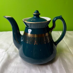 May include: Teapot with a teal glaze and gold accents. The teapot has a curved spout, a rounded body, and a handle. The lid is teal with a gold finial. Gold filigree decorates the body. The interior is white.