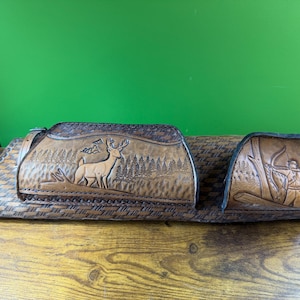 May include: A brown leather archery quiver set with a deer and archer design. The quiver set includes a main body with a deer scene and a smaller pouch with an archer scene. The set is made of brown leather and has a woven pattern.