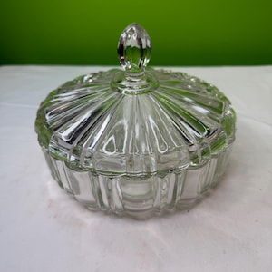 Op de afbeelding: Een heldere glazen snoepschaal met een deksel. De schaal is rond met een geribbeld ontwerp en een decoratieve knop aan de bovenkant. De schaal staat op een wit oppervlak met een groene achtergrond.