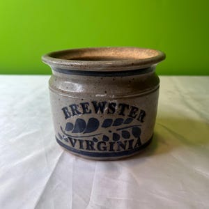 Puede incluir: Un pequeño tarro de gres gris con un diseño azul y blanco. El tarro tiene las palabras "Brewster Virginia" escritas en él.
