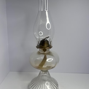 Puede incluir: Una lámpara de aceite de vidrio transparente con una base acanalada, una sección media bulbosa y una chimenea alta y cilíndrica. La lámpara tiene un quemador y una mecha de color latón. La lámpara está diseñada para contener aceite para la iluminación.