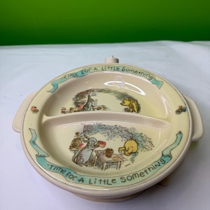 May include: A divided, cream-colored baby plate with illustrations of Winnie the Pooh characters. The plate has a suction base and handles. The text "Time for a Little Something" is written around the rim in a teal banner.