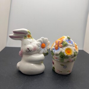 May include: A pair of ceramic Easter decorations. One is a white bunny holding a daisy, the other is a flower pot with colorful flowers. The bunny has pink accents and the flower pot has a floral design.