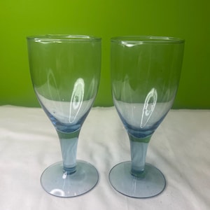 Puede incluir: Dos copas de vino de vidrio azul claro con una copa transparente y un tallo. Las copas están vacías y están sobre una superficie blanca.