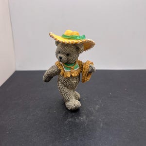 Peut inclure: Figurine d'ours en peluche gris portant un chapeau jaune et vert et un collier en forme de paille. L'ours tient un panier tressé. La figurine a une apparence texturée et détaillée et est placée sur un fond uni.