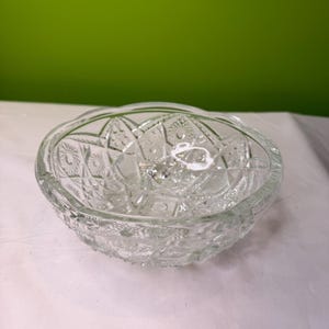 Puede incluir: Un cuenco de vidrio transparente con un patrón detallado y texturizado. El cuenco tiene un borde festoneado y una forma redonda. El intrincado diseño presenta elementos geométricos y florales, creando una estética vintage. El cuenco probablemente se utiliza para servir o exhibir.