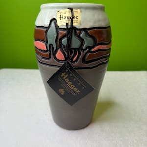 Könnte beinhalten: Eine Keramikvase mit grauem Sockel und cremefarbenem Oberteil. Der Mittelteil zeigt ein Landschaftsdesign in Braun, Grün und Koralle. Die Vase hat ein Etikett mit der Aufschrift "Royal Haeger, Made in USA."