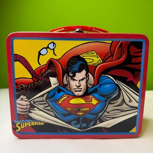 Può includere: Un portapranzo in metallo rosso con Superman. Il portapranzo ha uno sfondo giallo con un'illustrazione a fumetti di Superman nel suo costume. La parola "Superman" è stampata in basso.