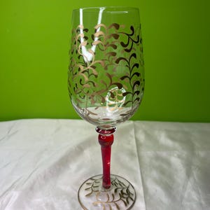 May include: A clear wine glass with a red stem and base. The glass is decorated with gold-colored, swirling leaf patterns. The base also features gold leaf designs. The background is a solid green color.