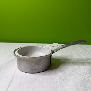 Puede incluir: Una pequeña cacerola plateada con un mango largo. La cacerola tiene un cuerpo redondeado y un borde alrededor de la parte superior. El mango está unido al lateral y se curva hacia arriba. La cacerola está sobre una superficie blanca.