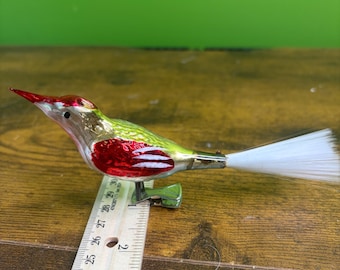 May include: A glass bird ornament with a red, green, and gold finish, featuring a long white fibre tail and a metal clip base. A wooden ruler is positioned underneath, showing the bird measures approximately 11.5 cm from beak to tail.