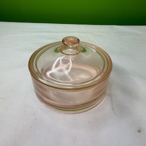 Könnte beinhalten: Ein klares, rosa Glasgefäß mit passendem Deckel. Das Gefäß ist rund mit einem kleinen Knopf auf dem Deckel. Das Gefäß steht auf einer weißen Oberfläche mit grünem Hintergrund.